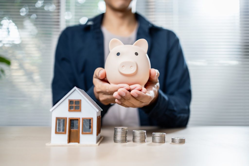 person holds piggy bank beside house model and coin stacks
